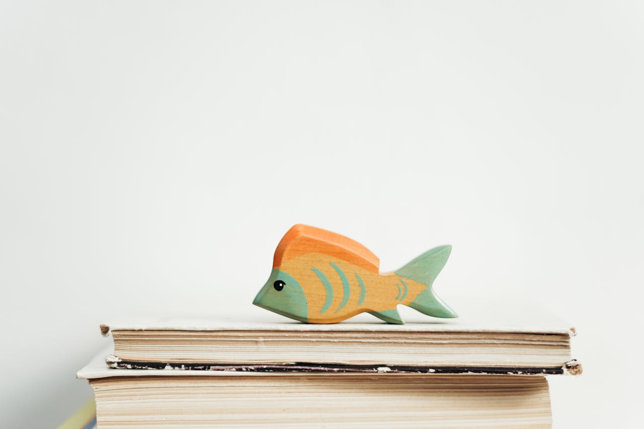 A colorful wooden fish toy sits atop a stack of vintage books against a plain background.